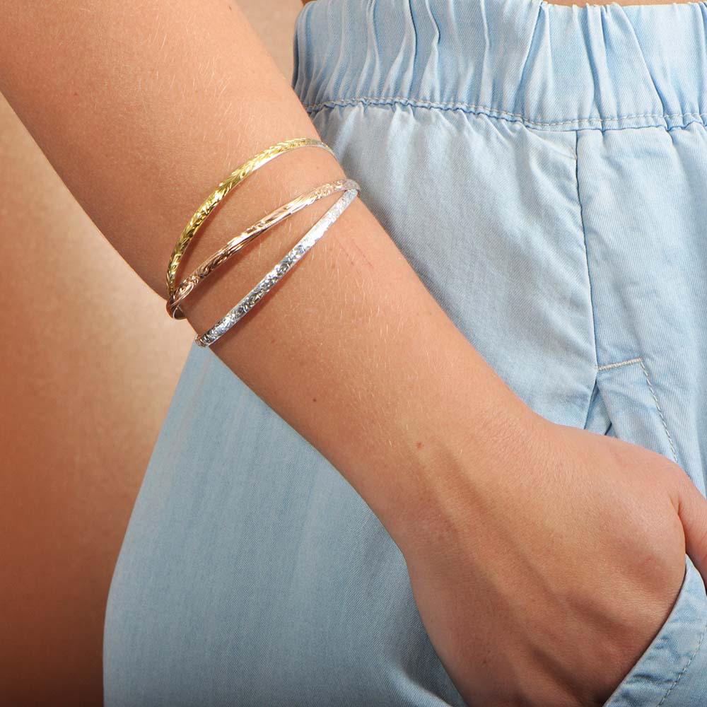 The photo shows a model wearing a set of three-tone gold vermeil Hawaiian scroll bangles.