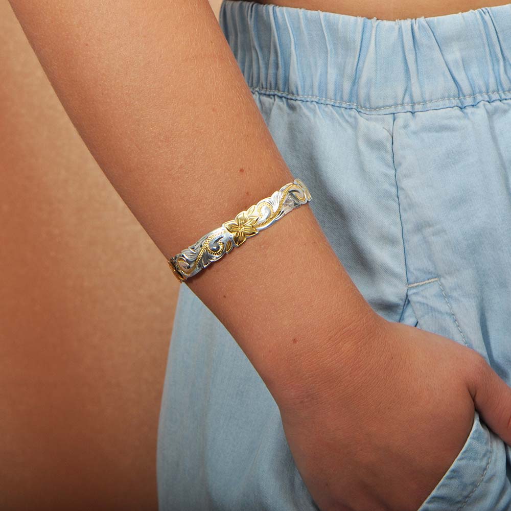 The photo shows the arm of a model wearing a two-tone white and yellow gold vermeil flowering plumeria cuff bangle. 