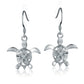 The photo shows a pair of white gold vermeil sea turtle hook earrings featuring a plumeria flower with a cubic zirconia gem. 